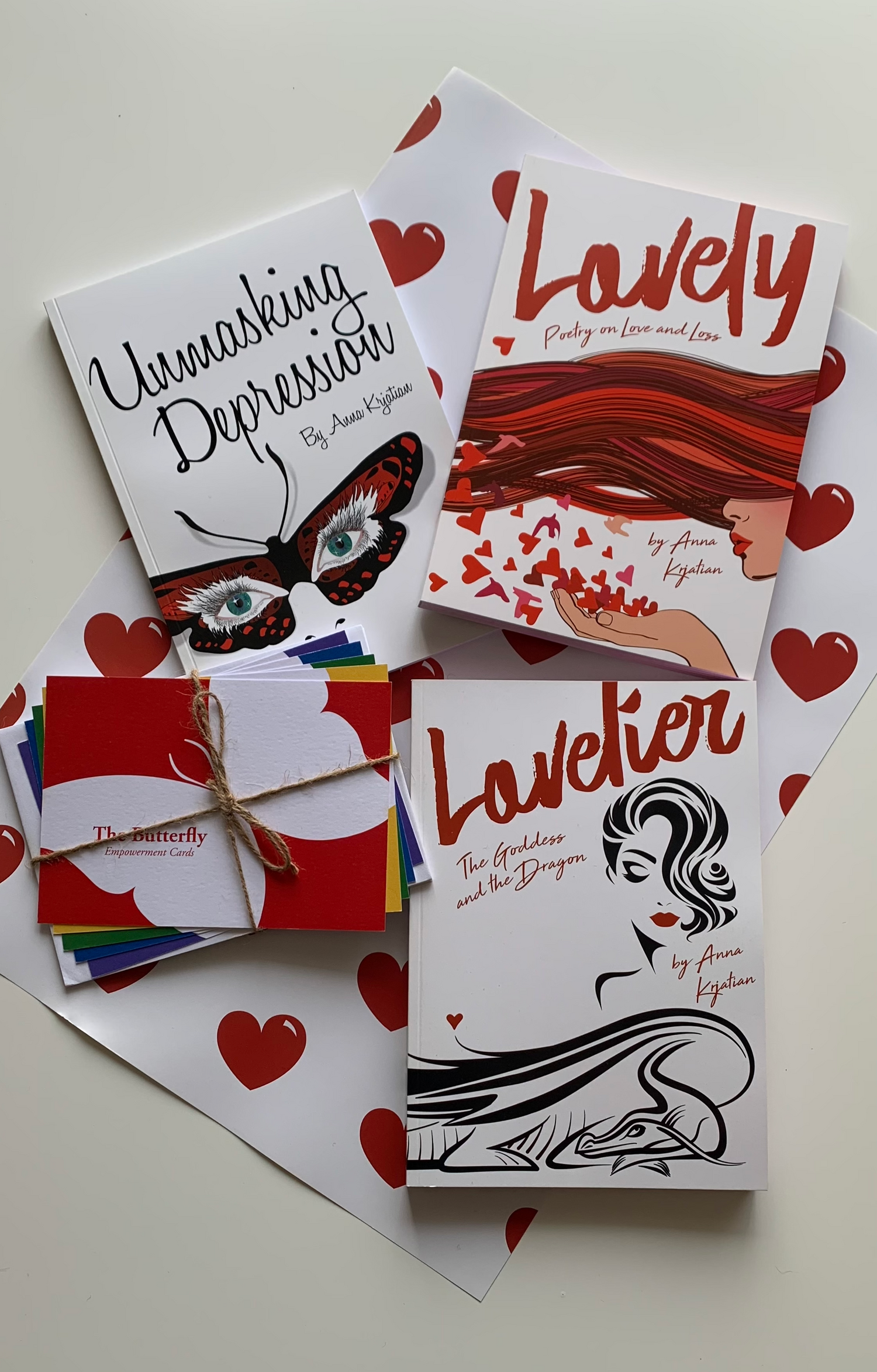 Three books "Unmasking Depression", "Lovely - Poetry on Love and Loss" and "Lovelier - The Goddess and The Dragon" sit on top of a white wrapping paper with bright red hearts on it. Next to the books is a pack of bright cards with a butterfly on them.