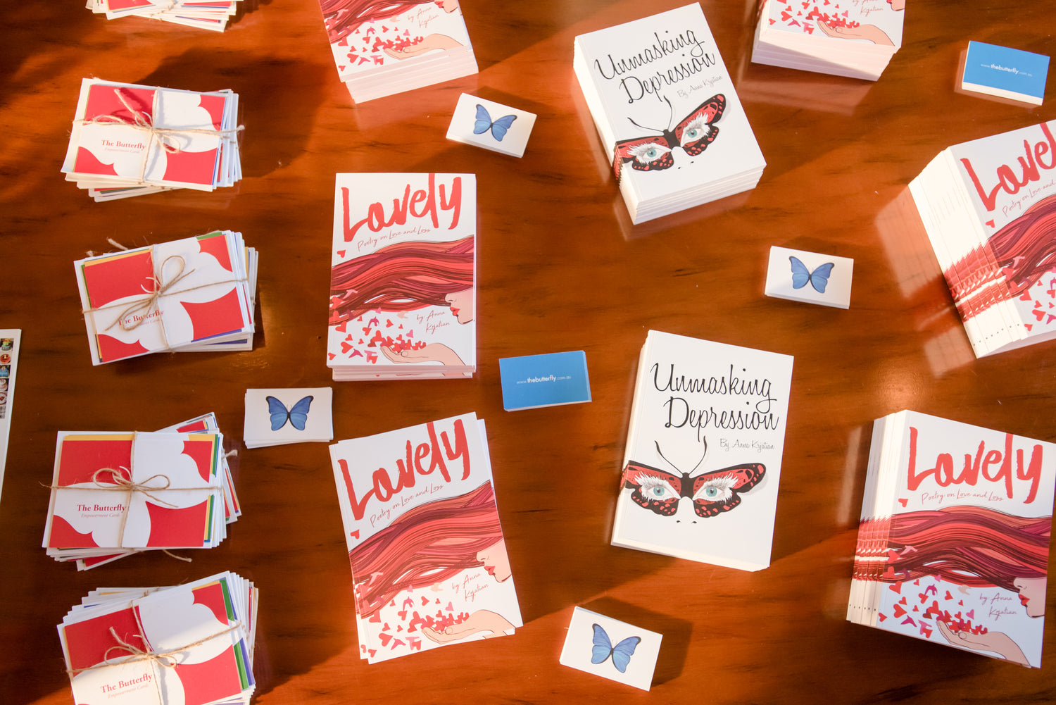 Red coloured Empowerment Cards and two books "Unmasking Depression" and "Lovely - Poetry on Love and Loss" and Butterfly business cards are set up on a brown table.