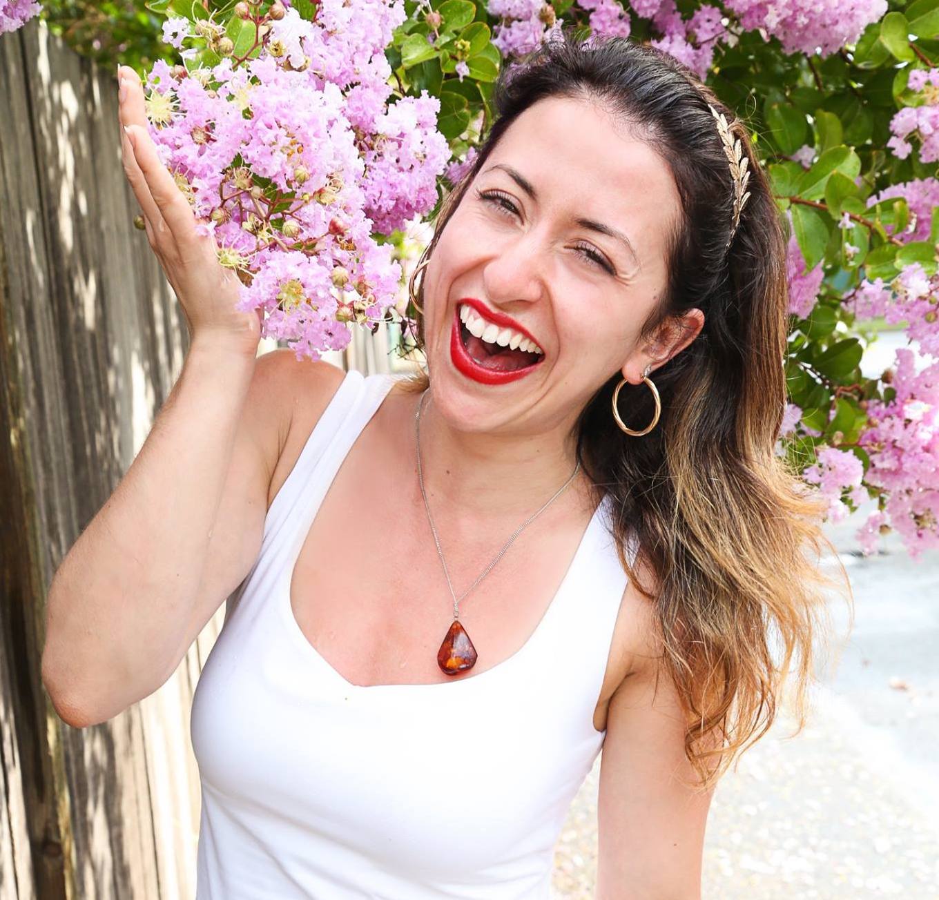 Anna Krjatian wears a white top, red lipstick, holding a pink flower in one hand, standing next to a fence and laughing.