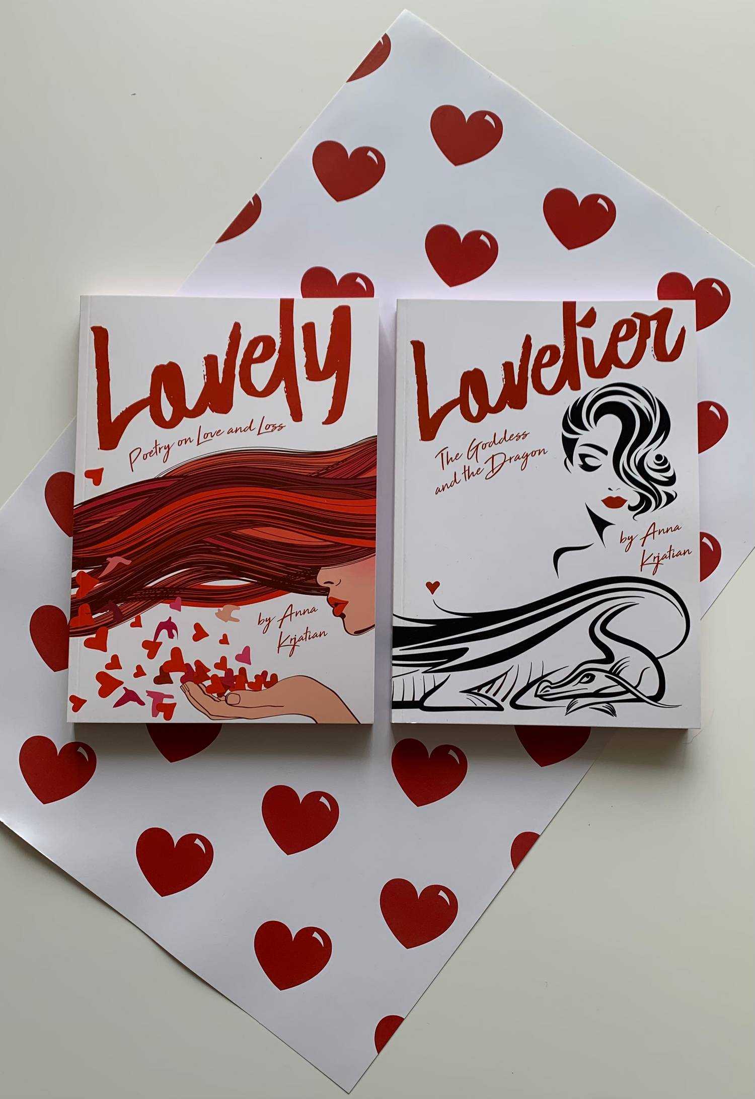Two books "Lovely - Poetry on Love and Loss" and "Lovelier - The Goddess and The Dragon" sit on top of a white wrapping paper with bright red hearts on it.