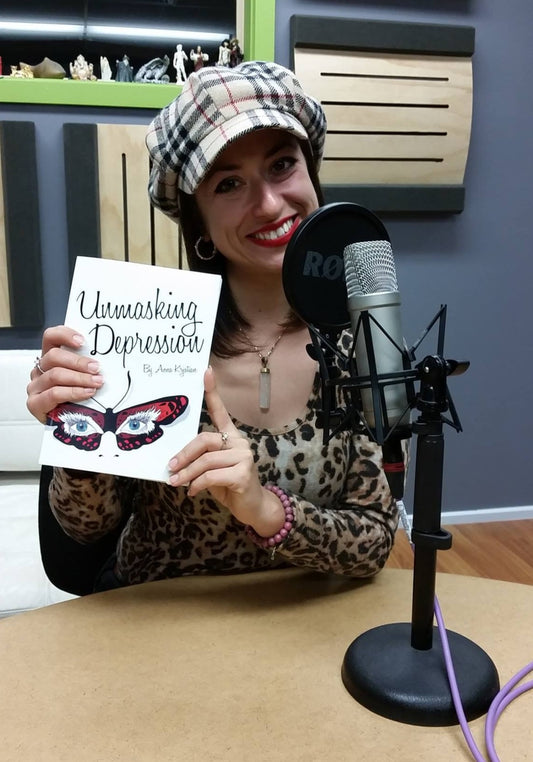 Anna Krjatian sits in front of a podcast microphone, wearing a hat and smiling at the camera and holding the book "Unmasking Depression."